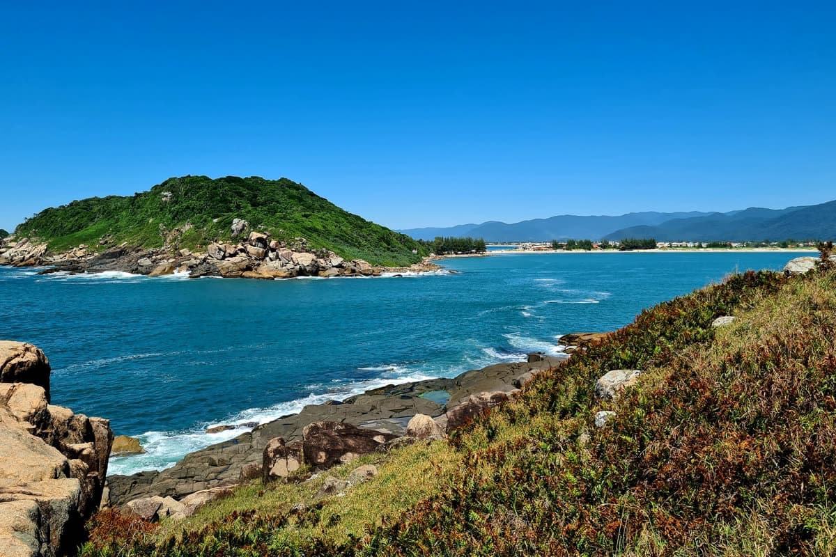A ilha verde da Ponta do Papagaio cercada de mar azul, vista do costão de pedras, com a praia e a serra ao fundo